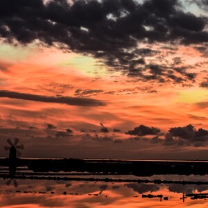 Può includere: Un tramonto drammatico sull'acqua, con mulini a vento in silhouette. Il cielo è una miscela di arancione e nuvole grigio scuro, riflessa nell'acqua calma. L'immagine cattura un'atmosfera pacifica e serena.