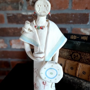 May include: A white ceramic figurine of a Native American figure, decorated with feathers, a beaded necklace, and earrings. The figure holds a small bowl and a drum with a blue and green design. The figure is wearing a white shawl with blue stripes.