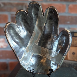 May include: A handcrafted, dark ceramic dish in the shape of a hand, with a glossy finish and gold accents. The dish is displayed on a black metal stand, showcasing its unique design and artistic craftsmanship. The background features a brick wall.