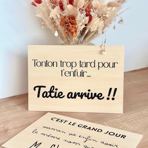 Puede incluir: Dos letreros de madera con texto negro. El letrero superior dice "Tonton trop tard pour t'enfuir... Tatie arrive !!" El letrero inferior dice "C'EST LE GRAND JOUR maman va enfin avoir le même nom que nous... Mme Chauvin !!" Un ramo de flores secas en el fondo.