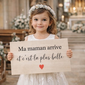 Puede incluir: Una niña con un vestido blanco y una corona de flores sostiene un letrero de madera con texto en francés y un corazón rojo. El letrero dice "Ma maman arrive et c'est la plus belle". El fondo muestra un interior de iglesia.