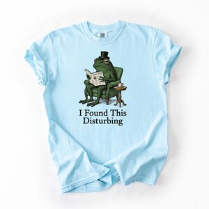 May include: Light blue t-shirt featuring a cartoon frog in a top hat, seated in a green armchair, reading a newspaper. The text "I Found This Disturbing" is printed below the frog illustration. The shirt is made of soft material.