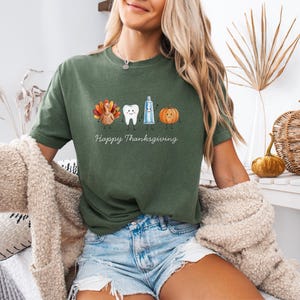 Op de afbeelding: Een olijfgroen t-shirt met korte mouwen en een afbeelding van een cartoonkalkoen, een tand, een tube tandpasta en een pompoen. Onder de figuren staat de tekst Happy Thanksgiving in een wit cursief lettertype. Het shirt heeft een ronde hals.