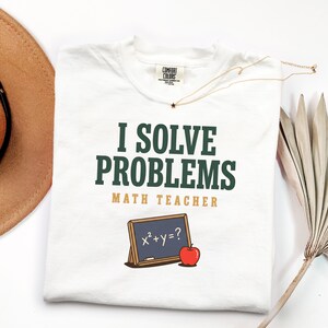 May include: White t-shirt with the text "I Solve Problems" in dark green, "Math Teacher" in gold, and a chalkboard graphic with an apple. A gold necklace with a star pendant is on the shirt.