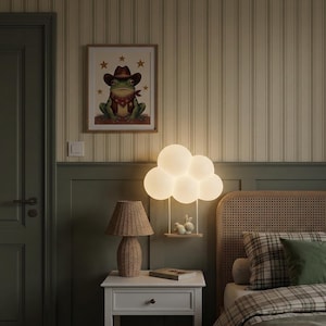Puede incluir: Un dormitorio con una ilustración enmarcada de una rana con sombrero de vaquero, una lámpara de pared en forma de nube, una lámpara de mesa de mimbre y una mesita de noche. La cama tiene un edredón a cuadros y una almohada verde. Las paredes están pintadas en tonos verdes y beige.