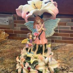 May include: A decorative figurine featuring two fairies standing among large, pale pink and white lily flowers. The larger fairy has light blue wings and a pink dress, while the smaller fairy wears a purple dress. A small ladybug rests on a petal.
