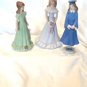 May include: Three porcelain figurines of young women. One in a green dress, one in a white dress holding a sign with "18", and one in a blue graduation gown holding a diploma. All are standing on a white surface.