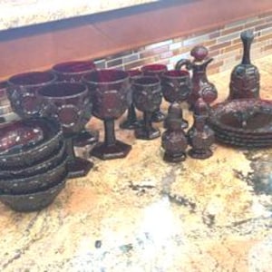 May include: A collection of dark red glass tableware including several stemmed goblets, small bowls, a pitcher, a bell, salt and pepper shakers, and a stack of plates. Each piece features a textured, embossed pattern across the deep red surface.