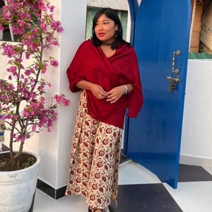 Puede incluir: Una persona con un chal rojo y una falda larga con estampado floral. La falda es de color crema con diseños florales rojos y marrones. El fondo incluye una puerta azul y una pared blanca con una planta en maceta.