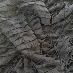 May include: Close-up of a dark gray fabric with a layered, ruffled design. The fabric has a textured appearance with horizontal rows of ruffles, creating a wave-like pattern. The material appears soft and flowing, with subtle variations in tone.