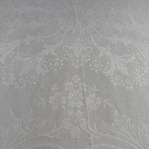 May include: A white tablecloth featuring a tone-on-tone damask weave. The fabric displays an intricate central medallion pattern composed of stylized floral motifs, leafy vines, and delicate petal clusters, creating a subtle textured surface.