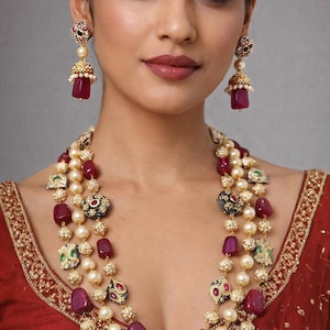 Op de afbeelding: Een vrouw draagt een gelaagde ketting en bijpassende oorbellen. De ketting heeft strengen van parels, rode edelstenen en sierlijke gouden accenten. De oorbellen hebben een vergelijkbaar ontwerp, met een rode edelsteen druppel.