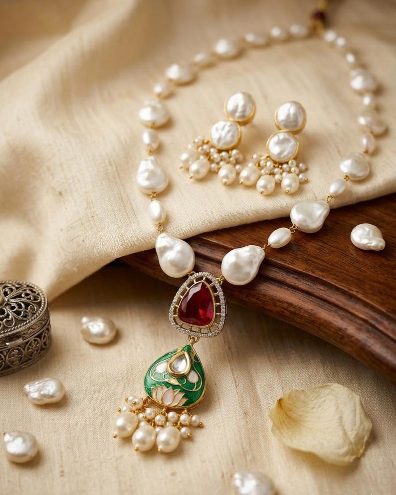 Puede incluir: Un conjunto de collar y pendientes con perlas blancas irregulares. El collar incluye un colgante central con una piedra roja grande, una forma de l&aacute;grima esmaltada en verde con un motivo de loto y peque&ntilde;os colgantes de perlas.