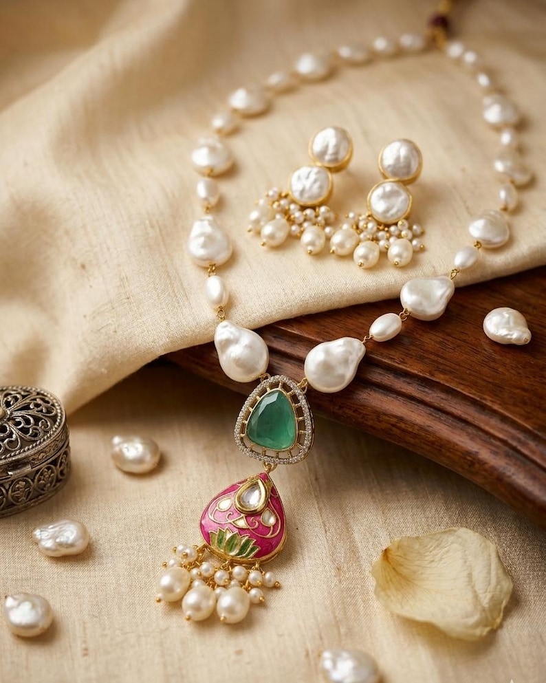 Puede incluir: Un conjunto de collar y pendientes con perlas barrocas blancas. El collar incluye un colgante central con una piedra verde triangular sobre una forma de l&aacute;grima esmaltada en rosa con un motivo de loto, acentuado con perlas blancas.