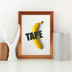 Può includere: Una stampa artistica incorniciata con una banana gialla con la parola "TAPE" in lettere nere. La stampa è in una cornice color rame, esposta su una superficie bianca con oggetti decorativi.