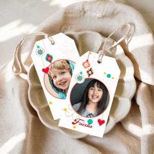 May include: Two white rectangular gift tags with rounded top corners and beige string loops. Each tag features a circular photo of a child, festive hanging ornament graphics in red, green, and teal, and swirling line accents. One tag displays the name Frieda.