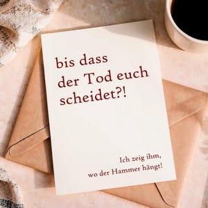 May include: A rectangular off-white greeting card featuring dark red text that reads: bis dass der Tod euch scheidet?! Ich zeig ihm, wo der Hammer hängt! The card is positioned on top of a light brown envelope, with a cup of dark coffee nearby.