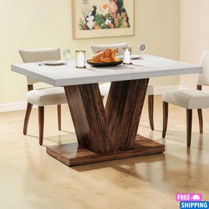 Puede incluir: Una mesa de comedor moderna con una superficie blanca y una base en forma de V de color marrón oscuro. La mesa está rodeada por cuatro sillas tapizadas de color beige. Un pollo asado y velas están sobre la mesa, lo que sugiere un entorno de comedor.