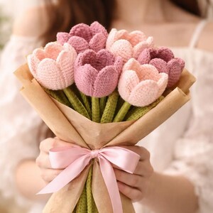 May include: A bouquet of handmade crochet tulips in shades of pink and purple, wrapped in brown paper and tied with a pink satin ribbon. The stems are green.