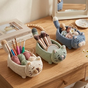 May include: Three handmade crochet cat-shaped baskets in cream, sage green, and blue. The cream basket holds yarn and crochet hooks, the green basket holds makeup brushes and a mirror, and the blue basket holds baby items.
