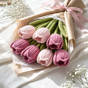May include: A bouquet of handmade crochet tulips in shades of pink and purple, with green stems, wrapped in brown paper and tied with a pink ribbon. The bouquet rests on a white surface, with a straw hat and white flowers in the background.
