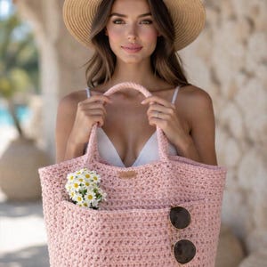 Puede incluir: Un bolso tote de ganchillo rosa claro con un bolsillo, que contiene un ramo de flores blancas y gafas de sol. El bolso tiene un asa rosa a juego. Se lleva un sombrero de paja.