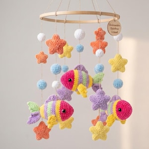 May include: A handmade crochet baby mobile featuring colorful fish, stars, and pom-poms. The mobile includes orange, yellow, purple, and blue elements, suspended from a wooden hoop. The text on the wooden tag reads "Underwater Rainbow School."