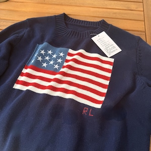 Könnte beinhalten: Marineblauer Pullover mit großem amerikanischem Flaggen-Design auf der Vorderseite. Die Flagge hat rote und weiße Streifen und ein blaues Feld mit weißen Sternen. Die Buchstaben "RL" sind rot gestickt. Ein weißes Etikett hängt am Pullover.