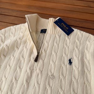Może przedstawiać: Kremowy sweter z warkoczowym splotem z zapięciem na zamek i prążkowanym kołnierzem. Na piersi małe granatowe logo gracza w polo. Zawiera metkę z logo "POLO RALPH LAUREN".