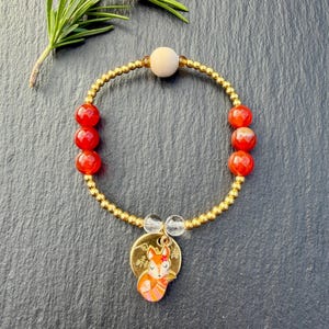 May include: A beaded bracelet featuring small metallic gold beads, six round red agate beads, one matte beige wooden bead, and two clear crystal beads. A circular gold-toned charm hangs from the bracelet, displaying an orange fox illustration.