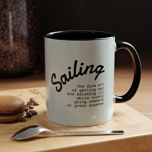 May include: A white ceramic mug with a black interior and handle. The mug features the word "Sailing" in a script font, along with a humorous quote about sailing. A spoon and a macaron are on a wooden surface.