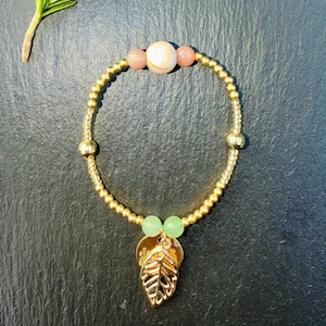 Peut inclure: Un bracelet de perles composé de petites perles métalliques dorées, deux perles sphériques vert clair, deux petites perles couleur pêche et une perle sphérique plus grande couleur crème. Une breloque feuille en métal doré est suspendue.