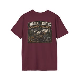 May include: A maroon short-sleeve t-shirt featuring a graphic print of a logging truck being loaded with timber in a forest setting. The text LOADIN' TRUCKS and Countin' DOLLARS with three dollar signs is printed above the illustration in a vintage font.