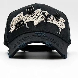Puede incluir: Gorra de béisbol negra con visera desgastada. La gorra presenta el texto "Backyard" en blanco, con una fuente texturizada y acentos negros. La gorra es de diseño clásico de seis paneles.