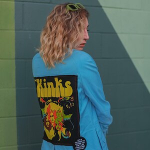 May include: A light blue jacket with a large patch on the back featuring the band "Kinks" and a colorful graphic. The jacket has a button on the front and a small pin with text. Green sunglasses rest on top.