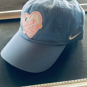 May include: A light blue baseball cap featuring a white Nike swoosh logo on the side and a heart-shaped patch on the front. The patch is pink and white with the text Cruel Summer and Miss You embroidered in yellow, blue, and pink thread.