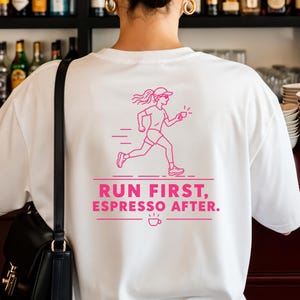Könnte beinhalten: Ein weißes T-Shirt mit einer pinkfarbenen Grafik einer laufenden Person und dem Text "RUN FIRST, ESPRESSO AFTER." sowie einem kleinen Kaffeetassen-Symbol. Das T-Shirt wird mit einer schwarzen Umhängetasche mit goldfarbenem Metallverschluss getragen.