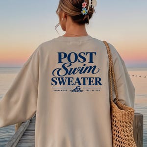 Könnte beinhalten: Ein beigefarbenes Sweatshirt mit Rundhalsausschnitt und dunkelblauem Text auf der Rückseite, der POST Swim SWEATER lautet, ergänzt durch eine kleine Grafik eines Schwimmers und die Worte SWIM MORE FEEL BETTER. Eine geflochtene Strohtasche.