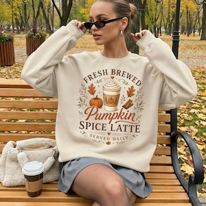 Könnte beinhalten: Ein cremefarbenes Langarm-Sweatshirt mit einem Grafikdruck einer Kaffeetasse, eines Kürbisses und Herbstblättern sowie dem Text "FRESH BREWED PUMPKIN SPICE LATTE SERVED DAILY" in brauner und orangefarbener Schrift auf der Vorderseite.