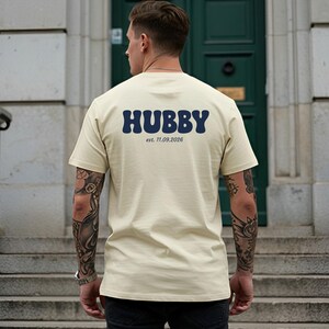 Puede incluir: Una camiseta de manga corta color crema con la palabra HUBBY en letras grandes, azul oscuro y estilo ondulado en la parte posterior. Debajo de la palabra HUBBY, el texto est. 11.09.2026 est&aacute; impreso en una fuente serif simple y azul oscuro.