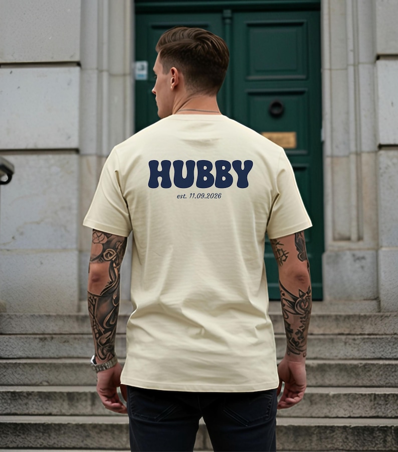 Puede incluir: Una camiseta de manga corta color crema con la palabra HUBBY en letras grandes, azul oscuro y estilo ondulado en la parte posterior. Debajo de la palabra HUBBY, el texto est. 11.09.2026 est&aacute; impreso en una fuente serif simple y azul oscuro.