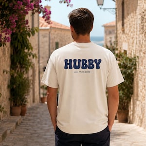 Puede incluir: Una camiseta de manga corta de color crema con la palabra HUBBY en letras grandes, azul oscuro y estilo ondulado en la espalda, con el texto est. 11.09.2026 impreso en una fuente m&aacute;s peque&ntilde;a y sencilla justo debajo.