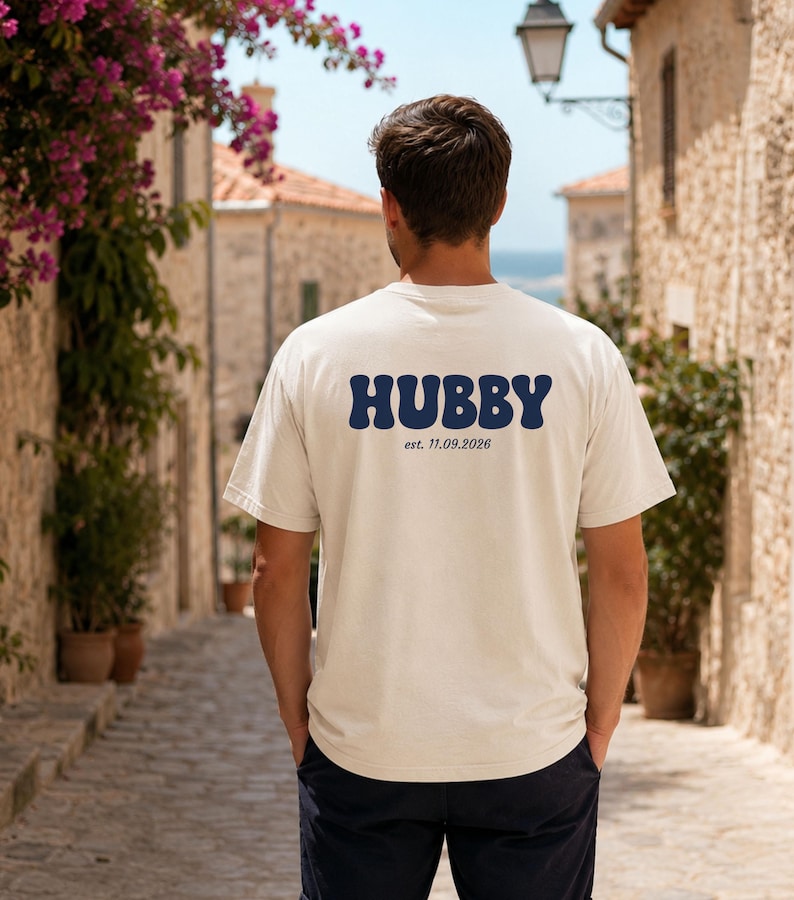 Puede incluir: Una camiseta de manga corta de color crema con la palabra HUBBY en letras grandes, azul oscuro y estilo ondulado en la espalda, con el texto est. 11.09.2026 impreso en una fuente m&aacute;s peque&ntilde;a y sencilla justo debajo.