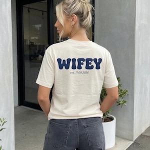 Puede incluir: Una camiseta de manga corta de color crema con el texto WIFEY en azul oscuro en una fuente ondulada y llamativa en la parte posterior, con el texto m&aacute;s peque&ntilde;o est. 11.09.2026 impreso debajo en una fuente serif negra simple.