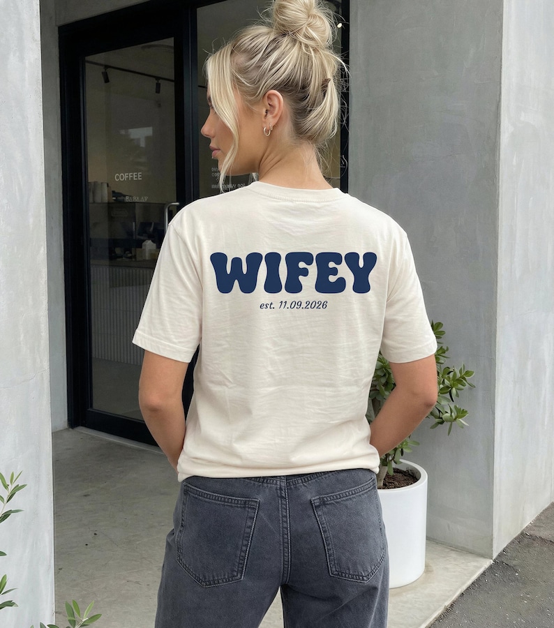 Puede incluir: Una camiseta de manga corta de color crema con el texto WIFEY en azul oscuro en una fuente ondulada y llamativa en la parte posterior, con el texto m&aacute;s peque&ntilde;o est. 11.09.2026 impreso debajo en una fuente serif negra simple.