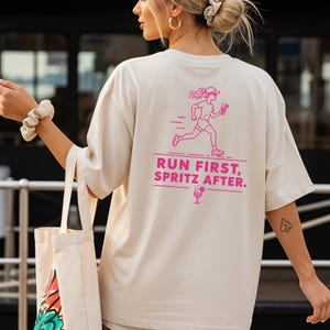 Könnte beinhalten: Ein beiges T-Shirt mit kurzen Ärmeln und einem pinkfarbenen Aufdruck auf der Rückseite. Das Motiv zeigt eine laufende Person mit einem Getränk in der Hand, darunter der Text "RUN FIRST, SPRITZ AFTER." und ein kleines Cocktailglas-Symbol.