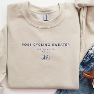 Könnte beinhalten: Ein beigefarbener Pullover mit Rundhalsausschnitt und dunkelblauem Aufdruck, der POST CYCLING SWEATER lautet, gefolgt von dem Text BETTER AFTER A RIDE und einem kleinen Fahrrad-Liniensymbol mittig auf der Brust. Das Kleidungsstück hat ein Gildan-Etikett.