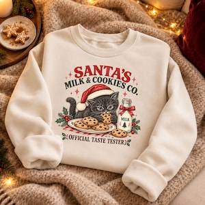 Könnte beinhalten: Ein cremefarbenes Sweatshirt mit Rundhalsausschnitt und einer Grafik eines schwarzen Kätzchens mit roter Weihnachtsmütze, das Schokoladenkekse neben einer Milchflasche isst. Der Text lautet SANTA'S MILK & COOKIES CO. OFFICIAL TASTE TESTER.