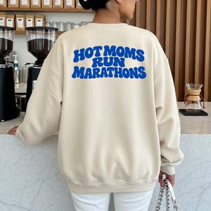 Könnte beinhalten: Ein beigefarbenes Sweatshirt mit Rundhalsausschnitt und dem Schriftzug "HOT MOMS RUN MARATHONS" in einer kräftigen, gewellten blauen Schrift auf der Rückseite. Das Kleidungsstück hat lange Ärmel, einen gerippten Ausschnitt und eine lockere Passform.