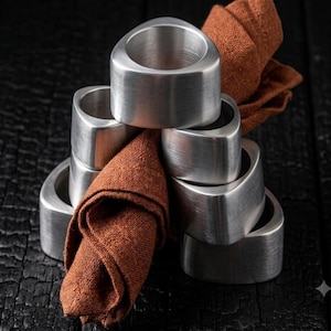 May include: A set of six brushed silver-tone metal napkin rings with a rounded triangular shape, stacked in a pyramid formation. A single rust-colored linen napkin is threaded through the center of the rings, creating a contrast in texture and color.